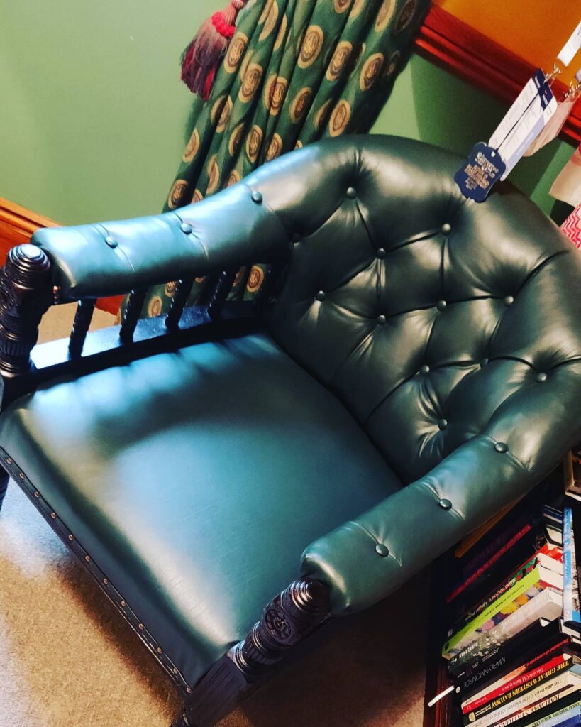 A dark green, tufted leather armchair with wooden armrests and carved legs sits beside a stack of colourful books. Restored by Winchester Furniture Repair, it stands out against green walls, a patterned curtain, and a hint of red trim. A blue tag hangs from the chair. Ryan Oliver Upholstery