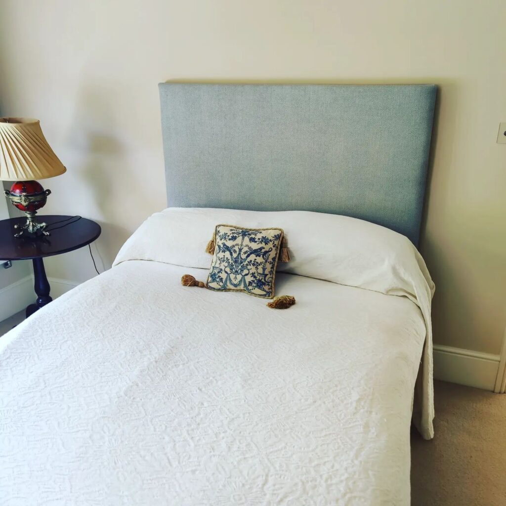 A neatly made bed with a light blue upholstered headboard, expertly crafted by Upholstery Winchester, white bedspread, and a decorative cushion with blue floral patterns and gold tassels. Beside the bed, a dark round bedside table holds a lamp with a pleated shade and red glass base. Ryan Oliver Upholstery