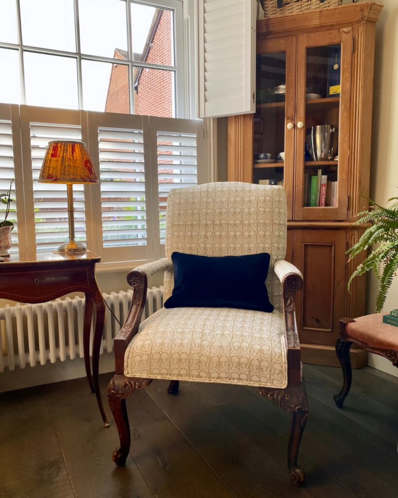 A classic armchair with patterned upholstery and a black velvet cushion sits on dark wood flooring by a window with white shutters. Nearby are a wooden cabinet with glass doors, a small side table with a stained glass lamp, and a fern plant. Ryan Oliver Upholstery Newbury
