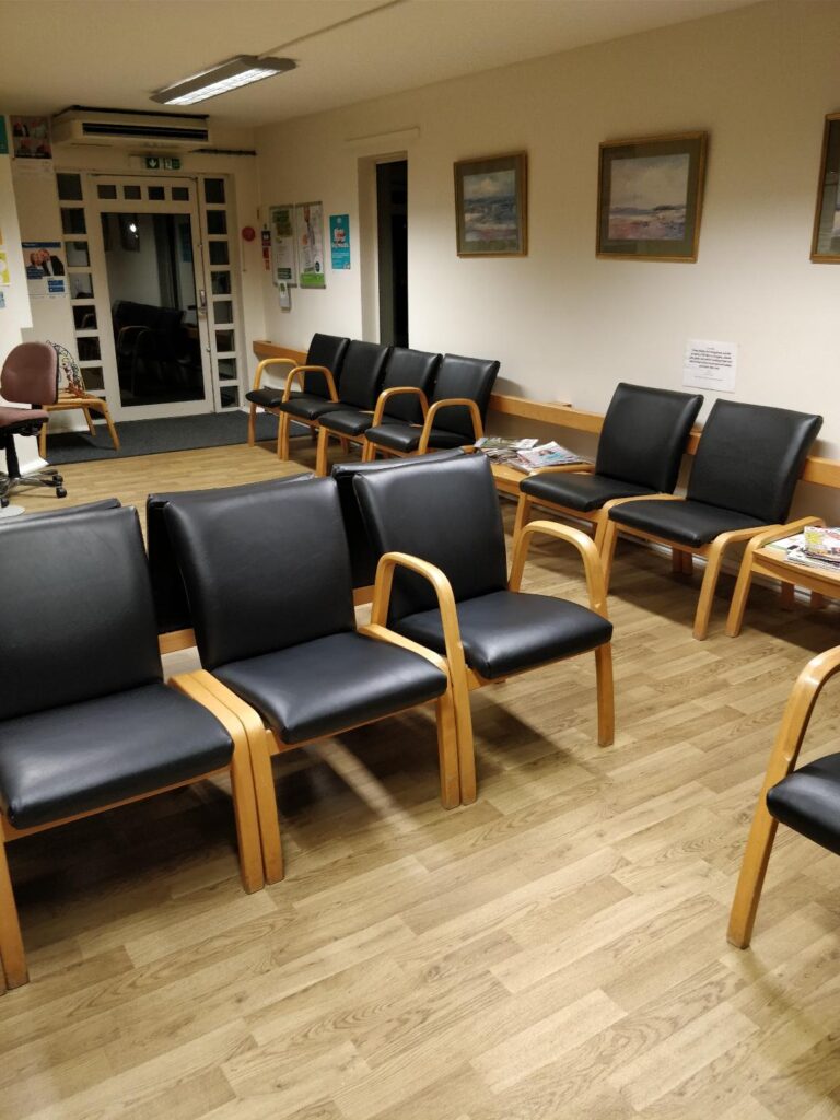 A doctor's waiting room with rows of empty black chairs with wooden frames—perfect candidates for Winchester Furniture Repair—light wood flooring, scattered magazines on side tables, framed pictures on cream walls, and a glass door at the back. Ryan Oliver Upholstery
