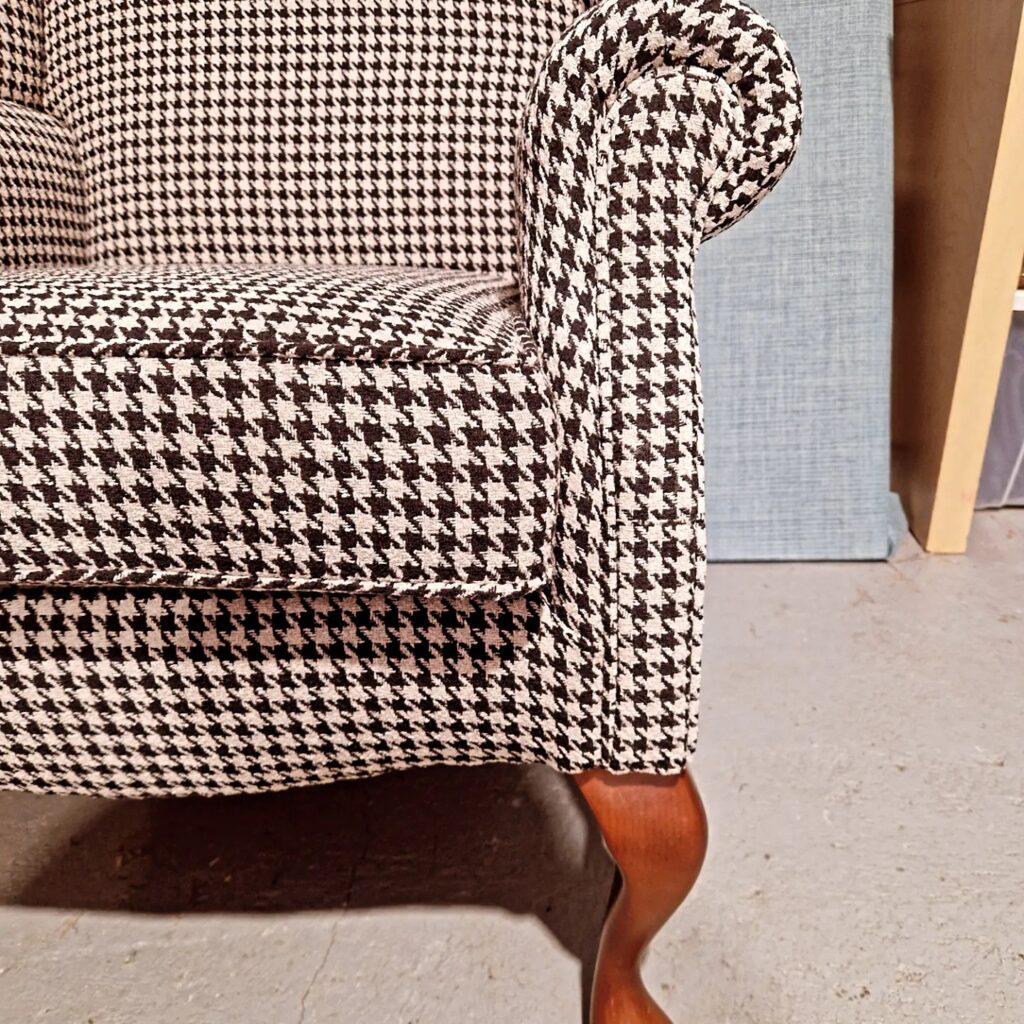 Close-up of a chair showing a rolled armrest and cushion upholstered in a black and white houndstooth pattern. The chair has a wooden cabriole leg and is placed on a grey concrete floor, with a textured panel and wood visible in the background. Ryan Oliver Upholstery
