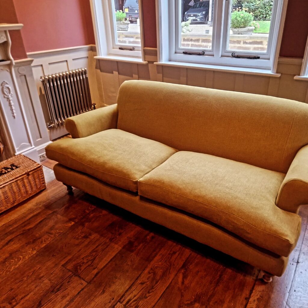 A mustard yellow upholstered sofa with curved arms, restored by Winchester Reupholstery, sits on a wooden floor in a living room with white panelling, a radiator, a wicker basket, and tall windows letting in natural light. Ryan Oliver Upholstery