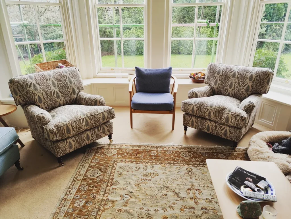 A bright sunroom with large windows features two patterned armchairs—perfect examples of Upholstery Winchester—facing inward, a blue-cushioned wooden chair, beige patterned rug, small table with magazines and a remote control, dog bed on the right, and lush garden views. Ryan Oliver Upholstery